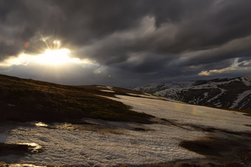 Mountain landscape. remnants of snow on the peaks, heavy dark clouds, the sun breaks through the clouds and illuminates the beautiful slopes, dry grass. Nice view of the sunset.
