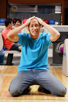 Man kneeling in bowling alley, hands on head, grimacing