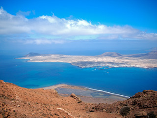 Sandy island with volcanoes and mountains in Atlantic Ocean. Aerial view from mountain slope. Yacht harbor and boats in strait. Deep blue sky with white clouds. La Graciosa, Canary Islands, Spain