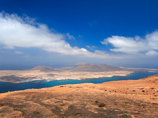 Sandy island with volcanoes and mountains in Atlantic Ocean. Aerial view from mountain slope. Yacht harbor and boats in strait. Deep blue sky with white clouds. La Graciosa, Canary Islands, Spain