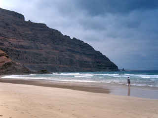 Lonely photographer on a sandy ocean beach shoots the surf under a rock slope of the volcano. Deep dark blue sky with clouds. Lanzarote, Canary Islands, Spain