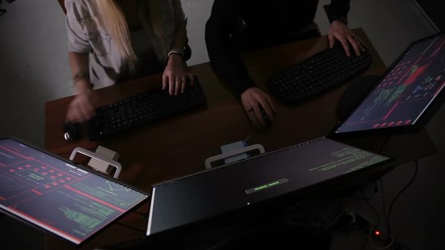 Male and female hackers working on computers with data code on display screens in a dark room. HD.