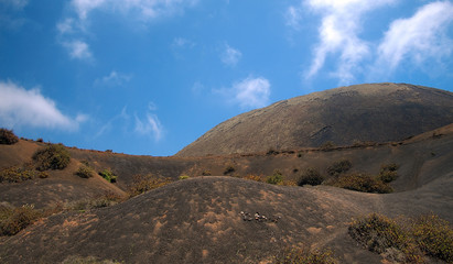 Mountain volcanic hillsides with rare plants on the background of deep blue sky with white clouds. Lanzarote, Canary Islands, Spain