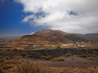 Fototapeta na wymiar Mountain valley with white buildings and villages between old volcanic slopes. Red Earth and green fields on the background of deep blue sky with white clouds. Lanzarote, Canary Islands, Spain