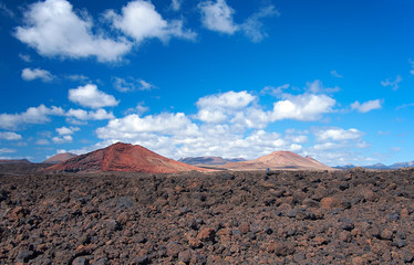 Red volcanoes and mountains on the background of deep blue sky with white clouds. Field of black volcanic bombs in the foreground. Lanzarote, Canary Islands, Spain
