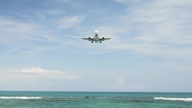 Airplane landing on island Bali airport under blue sea with waves on the horizon. Cinematic shot of overhead flying aircraft with beautiful ocean on cloudy sky background. Focal length 50 mm