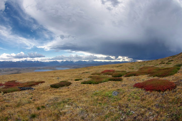 A large storm cloud on the alpine steppe of the yellow dry grass and lake with high snow peaks range on the horizon Plateau Ukok, Altai mountains Siberia, Russia