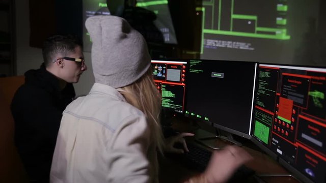 Male and female hackers working on computers with data code on display screens in a dark room. HD.
