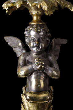 Praying brass angel on black background. A winged putto made of brass, covered with silver, as part of a candelabra from the nineteenth century and a symbol for religious passion. Macro object photo.