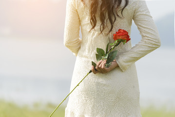 Beautiful women wearing white dress standing behind holding a red rose.A woman looking at the lake in front of her.