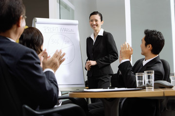 Businesswoman standing next to flipchart, other executives clapping