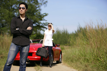 Man standing with arms crossed, woman and red sports car in the background