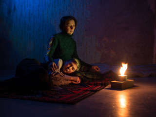 Fototapeta premium Adult woman sitting on the floor and sadly looks and the young boy sleeping on her lap against the backdrop of concrete walls with torn wallpaper and a burning candle