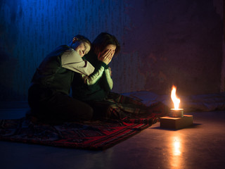 Adult woman sitting on the floor and crying and the young boy put his head on the shoulder of his mother against the backdrop of concrete walls with torn wallpaper and a burning candle