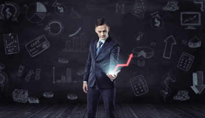 Businessman holding computer projection of rising red arrow on the background with economic sketches.