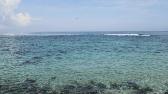 Airplane landing on island Bali airport under blue sea with waves on the horizon. Cinematic shot of overhead flying aircraft with beautiful ocean on cloudy sky background. Focal length 28mm