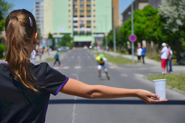 Young girl hold cup of water during roller skating competition and helping the athletes to hydrate...
