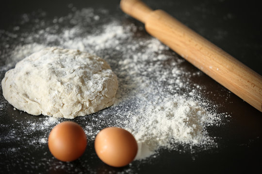 Dough with flour, eggs and rolling pin on dark table