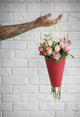 Hand of tattooed man holding paper cone with beautiful bouquet against brick wall