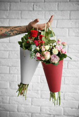Hand of tattooed man holding paper cones with beautiful bouquets against brick wall