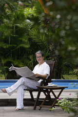 Mature man sitting by swimming pool, reading newspaper