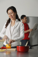 Mature woman in kitchen preparing a meal, man in background holding newspaper