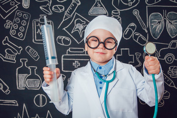 Happy little boy in doctor costum holding syringe and sthetoscope on dark background with pattern. The child has glasses