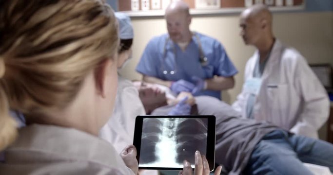 Young woman doctor looks at X-ray images on a tablet computer while casualty ward staff look after injured patient in background. Recorded hand-held.