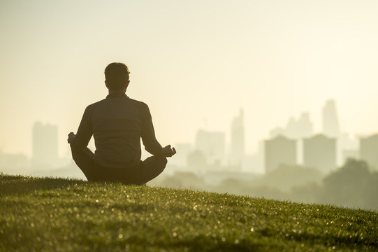 Silhouette of a man sitting in the lotus position meditating on the grassy top of Primrose Hill in front of a misty golden sunrise view of the London city skyline