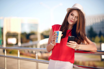 Happy and young beautiful girl in a fashionable hat, red shirt and nice long hair drinking coffee outdoors in the summer in the city and looking at the phone