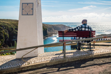 Lonely old man sitting and looking at the beautiful landscape with ocean and cliffs in Alentejo, Portugal