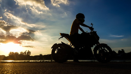 Silhouette biker with his motorbike beside the natural lake and beautiful evening light.