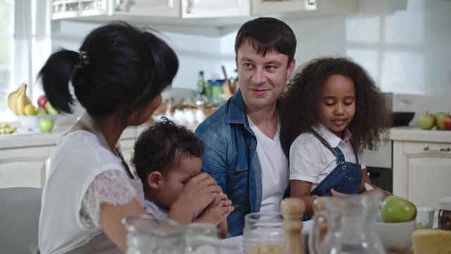 Parents sitting in the kitchen, having breakfast with their little kids on laps, cute curly daughter playing with kitchen stuff as mom holding a glass of milk and feeding her baby son 