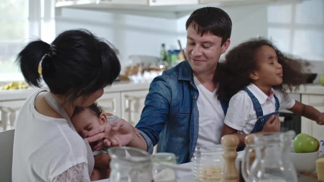Slow motion of a father wiping milk from face of his little son in the kitchen, cute daughter sitting on lap of her dad, shaking her head with her thick curly hair flying