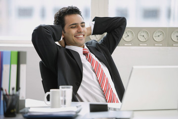 Businessman sitting in office, hands behind head, smiling