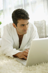 Man in bathrobe, lying on floor at home, using laptop