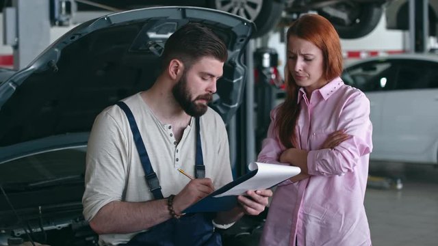 Young auto mechanic standing near car with opened hood and discussing something with female customer