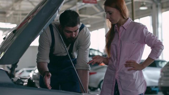 Professional mechanic checking engine at repair shop and listening to female client pointing at broken details under the hood of car