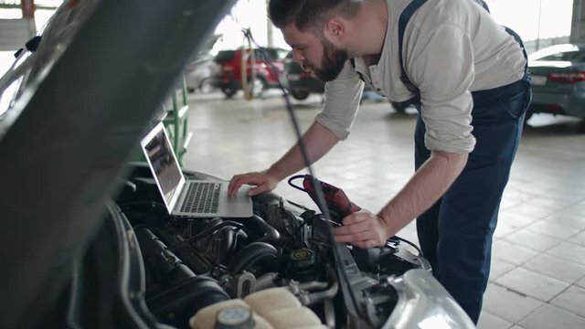 Tilt up of auto service worker examining details under the hood of car and using laptop 