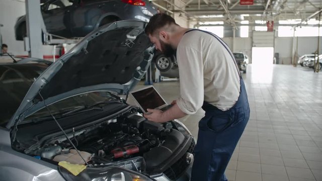 Technician walking to car in the repair garage and placing laptop under the hood for auto diagnostics