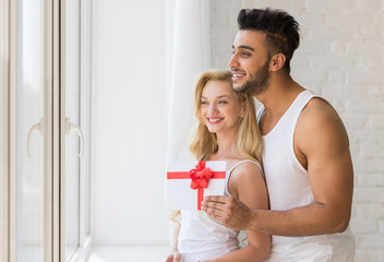 Young Beautiful Couple Stand Near Big Window, Hispanic Man Give Woman Present Envelope With Ribbon, Happy Smile Lovers