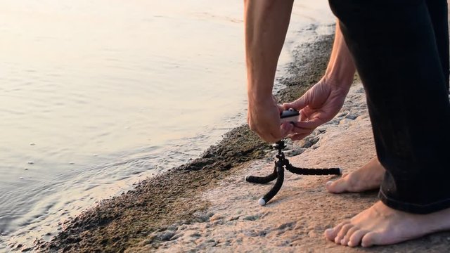 Barefooted man installs white action camera in waterproof case on tripod on sea shore