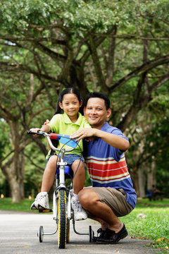 Girl on bicycle, father crouching down next to her, both looking at camera