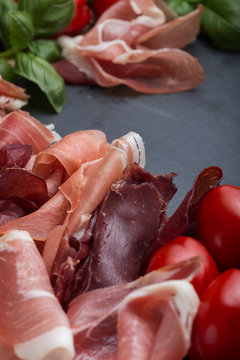 Ham mix. Jamon. Traditional Italian and Spanish salting, smoking, dry-cured dish - jambon Serrano and prosciutto crudo sliced with herbs and tomatoes on dark stone background. Copy space. Closeup. 