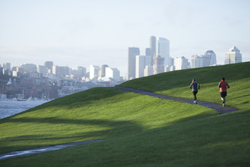 An adult man and woman running the paved path through Gasworks Park with the Seattle cityscape in the background. Washington State.