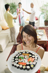 Woman holding up plate of sushi, smiling at camera, people in the background