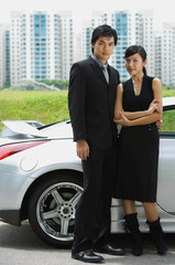 Couple standing side by side, silver sports car behind them
