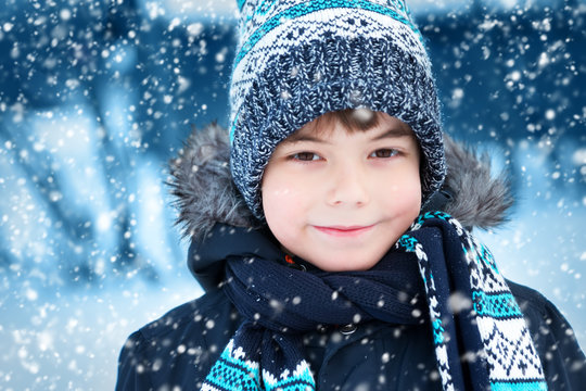 Boy in knitted hat, gloves and scarf outdoors at snowfall. Child in winter clothes