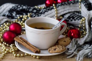 Obraz na płótnie Canvas Horizontal photo of hot green tea in a white mug with spoon, cinnamon sticks, chocolate cookies, knitted blanket and christmas decoration