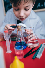 Close up of happy little scientist looking colorful chemical liquid in flask through magnifier against of drawn blackboard